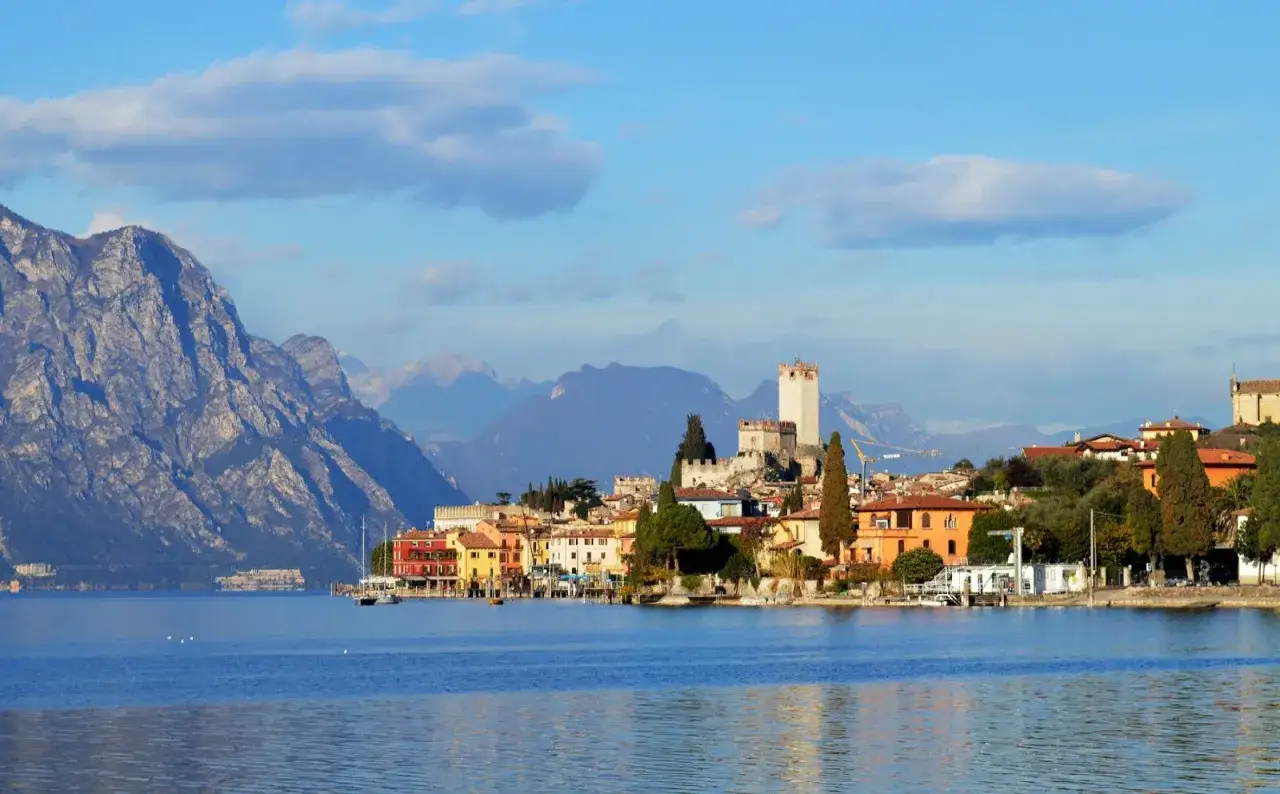 Paesi del lago di Garda sponda veronese: un borgo con castello domina la riva, circondato da montagne imponenti.