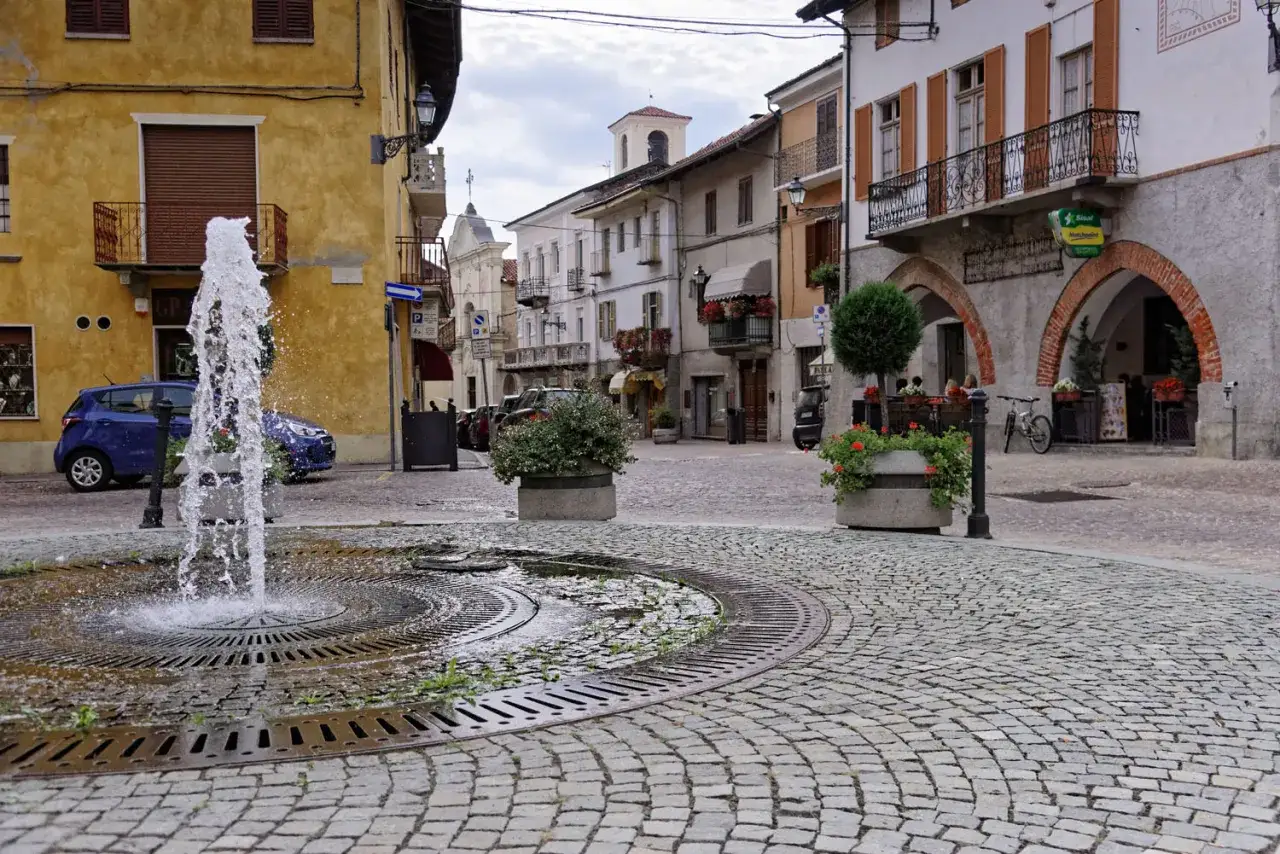 Fontana zraszająca wodę na brukowanym placu w San Giorgio Canavese, otoczonym kolorowymi budynkami i zielenią.