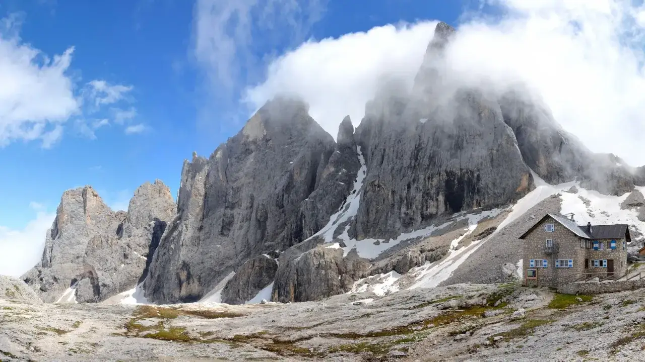 Rifugio in pietra ai piedi di imponenti vette rocciose, con neve residua e cielo azzurro. Un luogo incantevole al passo Valles.