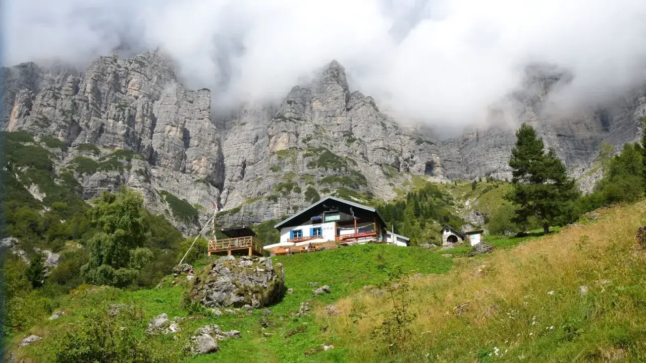 Rifugio alpino tra imponenti pareti rocciose delle Dolomiti Bellunesi, immerso nel verde.