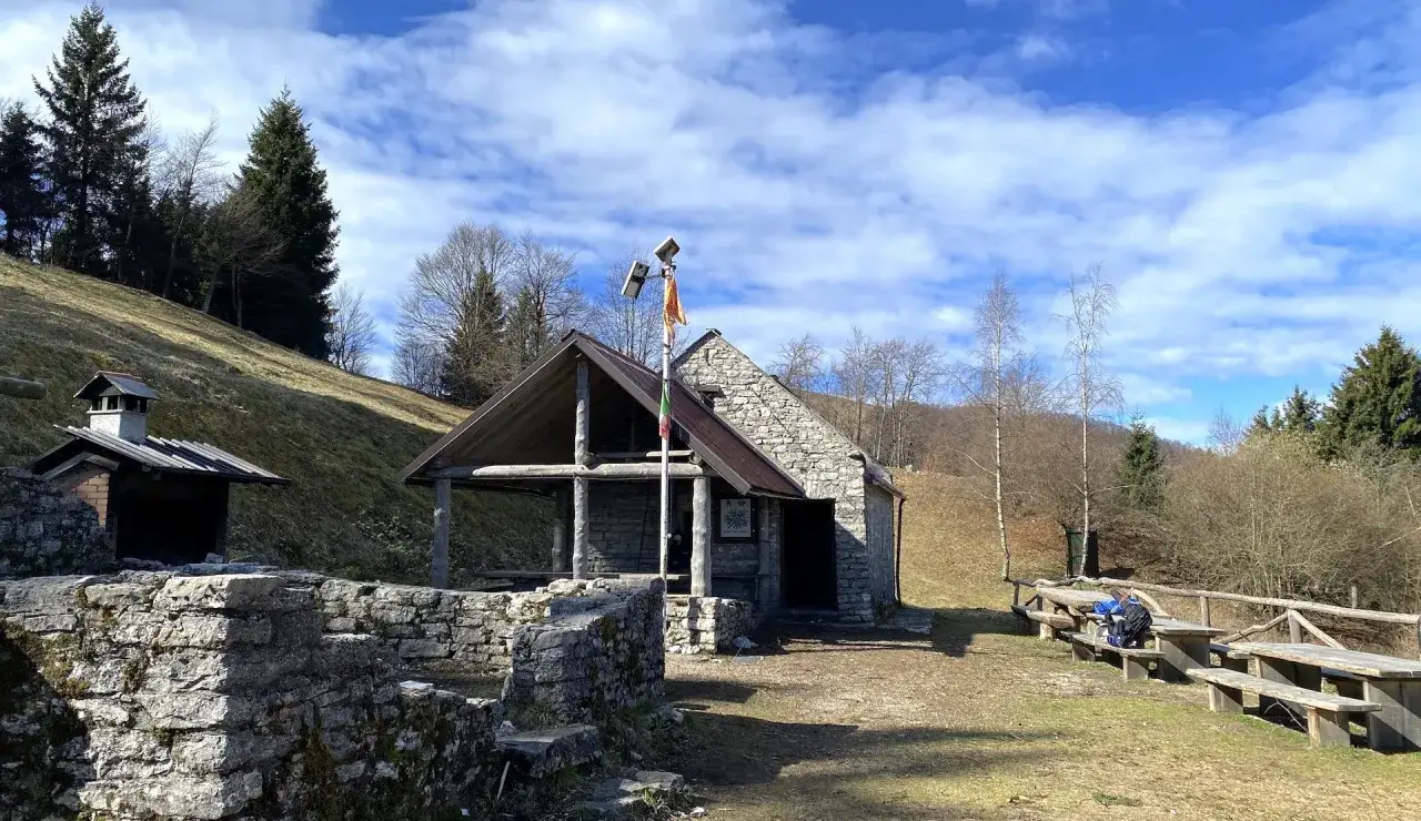 Bivacco Vallon Scuro: rifugio in pietra con tetto spiovente, tavoli in legno e barbecue in muratura, immerso nel verde di un pendio boscoso.