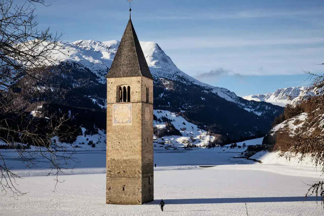 Torre sommersa nel lago di Resia ghiacciato, con montagne innevate sullo sfondo. Un uomo cammina sulla neve.