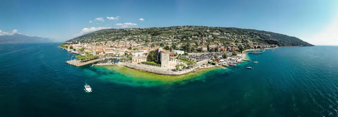 Panoramica di Torri del Benaco con il suo castello, il porto e la spiaggia baciata dal sole. Acque turchesi invitano a un tuffo.