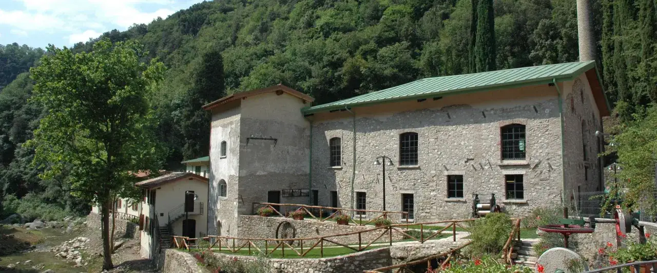 Antica cartiera a Toscolano Maderno, cosa vedere: un complesso edilizio in pietra con tetto verde, immerso nel verde, con un ruscello e un grande albero.