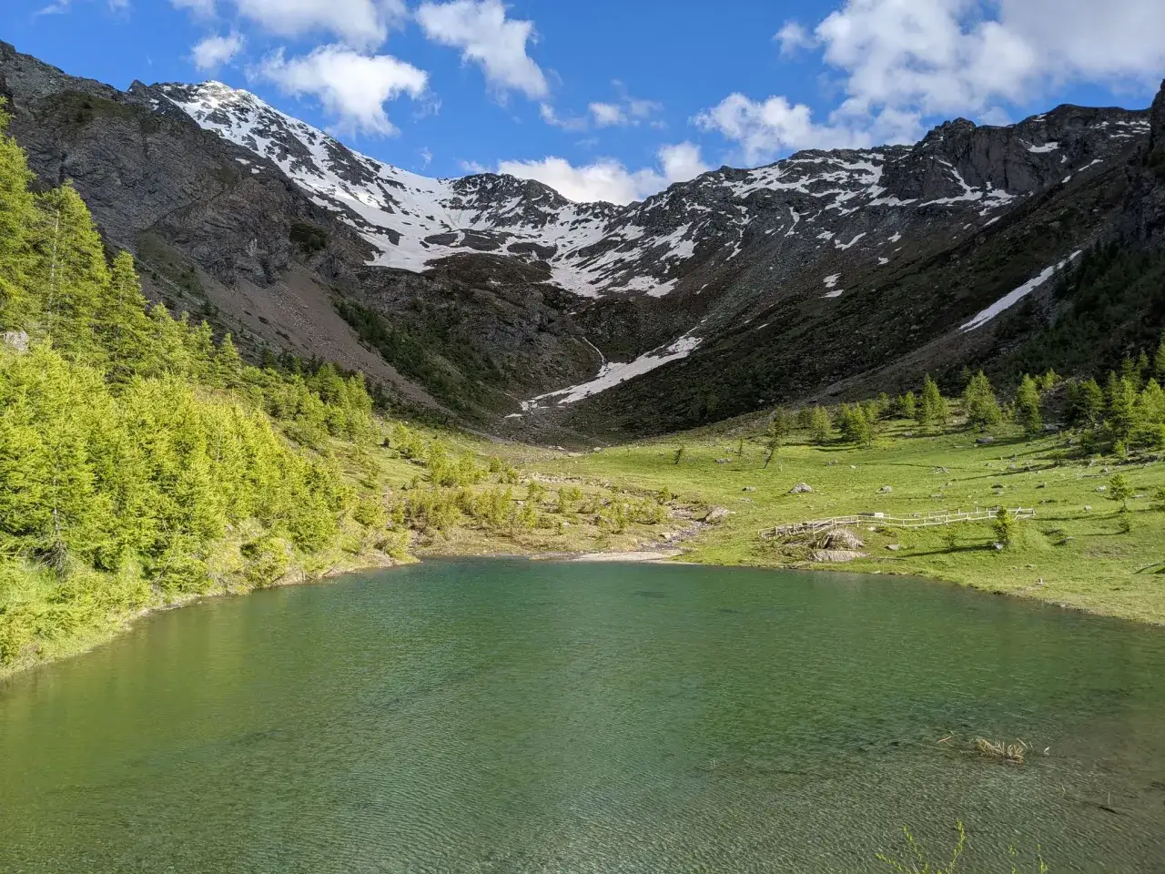 Sestriere estate: un lago alpino incastonato tra montagne innevate e verdi pascoli, con un cielo azzurro punteggiato di nuvole.