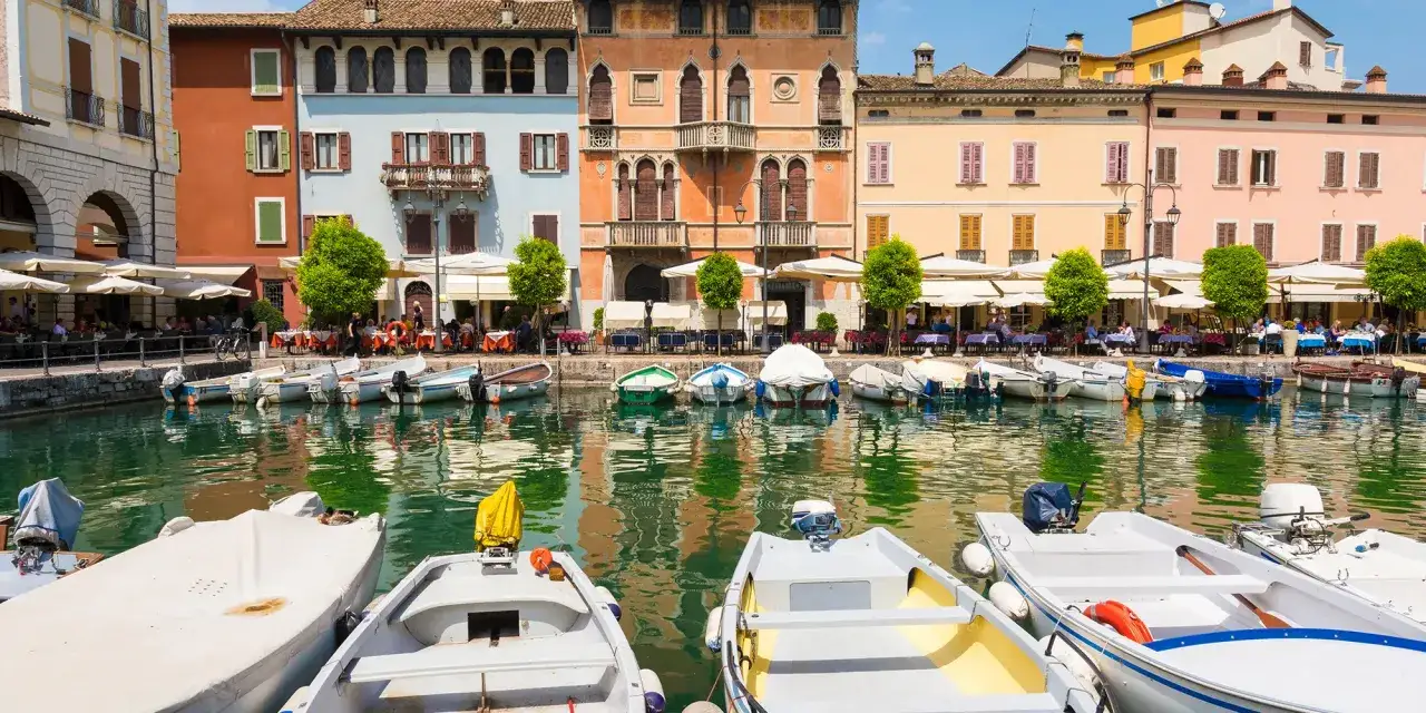 Barche ormeggiate nel porto di Moniga del Garda, con case colorate e tavolini all'aperto che si riflettono sull'acqua.