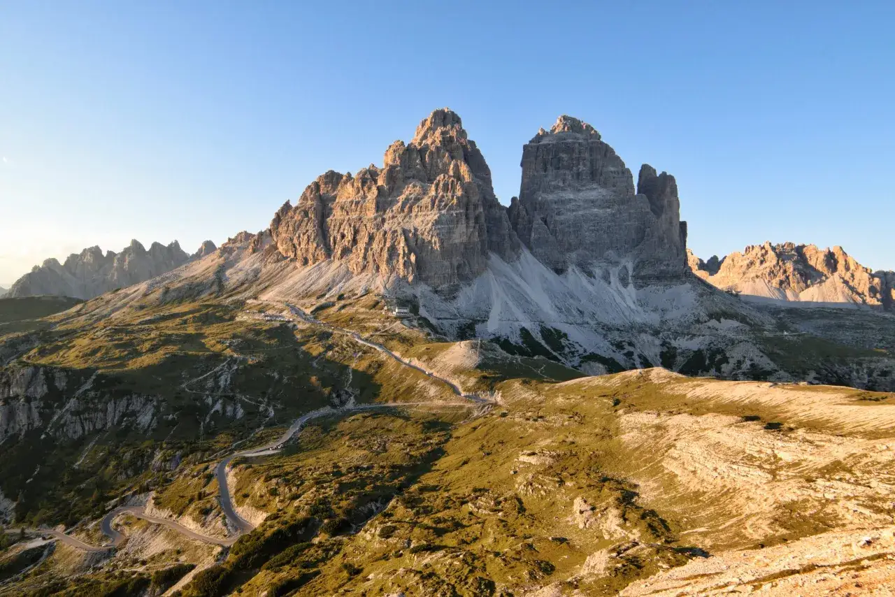 Le maestose Tre Cime di Lavaredo, icona dei Cadini di Misurina, si stagliano contro un cielo azzurro. Un sentiero serpeggia tra i prati verdi e le rocce.