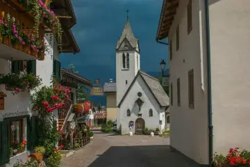 Un pittoresco vicolo a Rocca Pietore, con case fiorite e una chiesa dal cielo tempestoso.