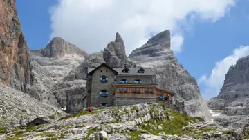 Il rifugio Tuckett e Sella, un'oasi di pietra tra le imponenti vette dolomitiche, accoglie escursionisti sotto un cielo azzurro e nuvoloso.