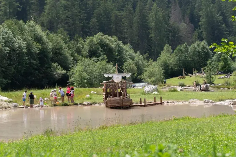 Bambini giocano vicino a un galeone di legno in un lago, mentre altri si rilassano sull'erba. Perfetto per le **san vigilio di marebbe passeggiate** estive.