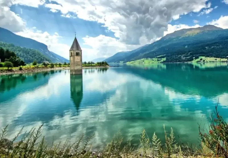 Torre sommersa nel lago di Resia, con montagne verdi e cielo nuvoloso.