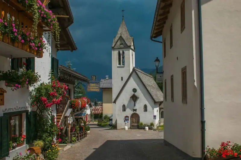 Un pittoresco vicolo a Rocca Pietore, con case fiorite e una chiesa dal cielo tempestoso.
