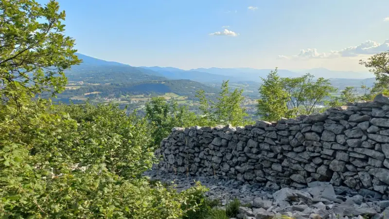 Muro di pietra a secco con vista panoramica su una valle alpina e montagne all'orizzonte.