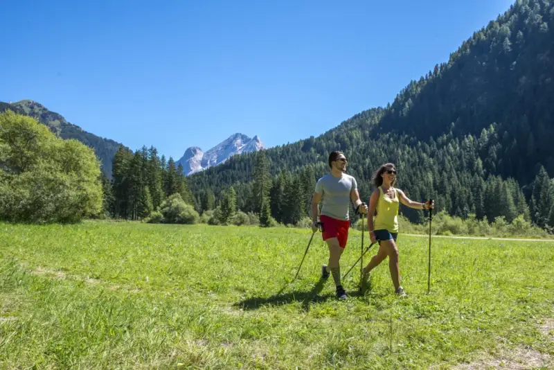 Coppia fa nordic walking in un prato verde con montagne sullo sfondo, ideale per canazei escursioni facili.